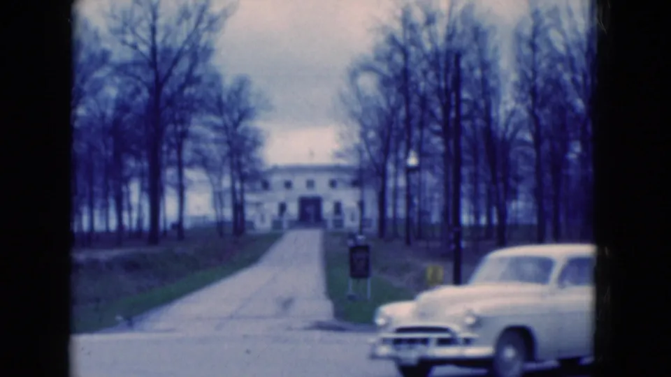 1975 Florida 8mm frame showing a white period car crossing the foreground before the driveway and large white building.