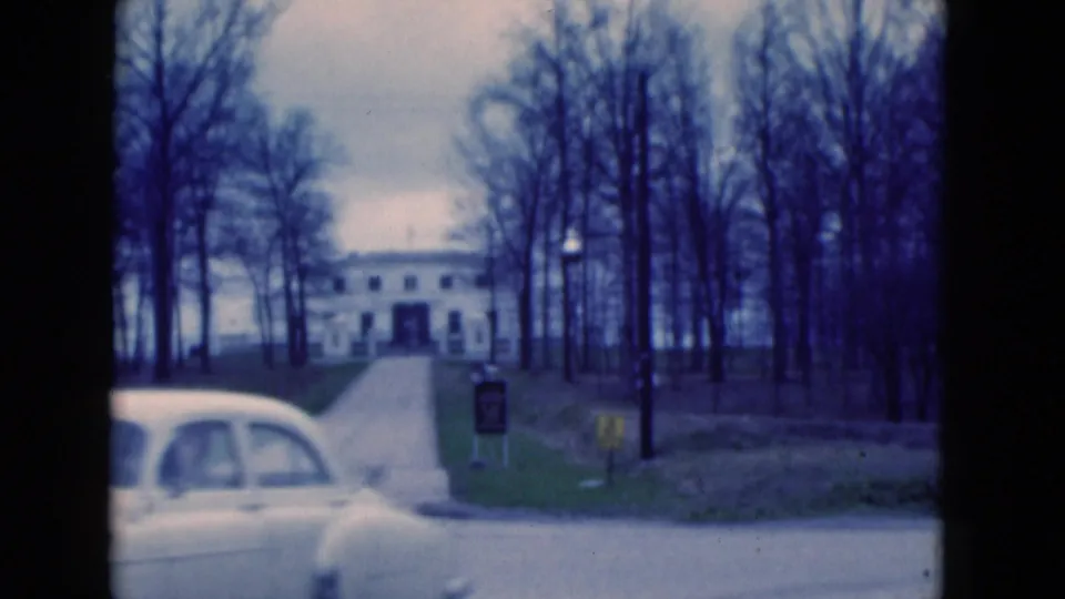 1975 Florida home-movie frame with a white car partly blocking the driveway view toward a large white building among bare trees.