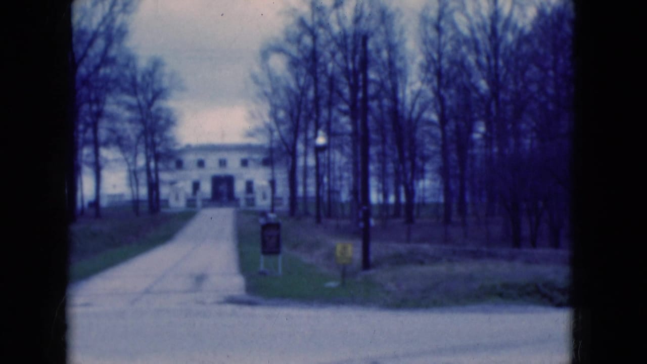 White building at the end of a paved driveway, framed by bare trees under an overcast sky.