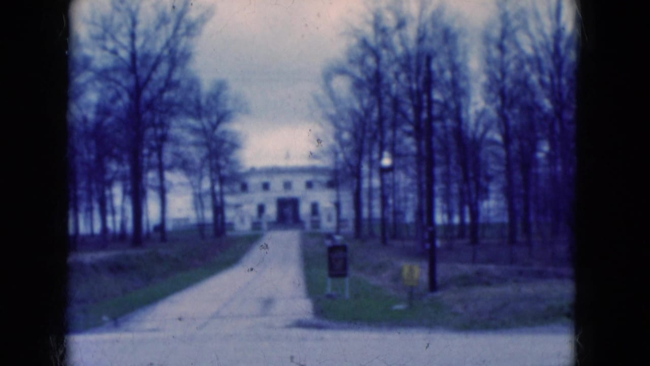 1975 Florida 8mm frame of a white building approached by a driveway, with leafless trees, lawn, and signs near the foreground road.