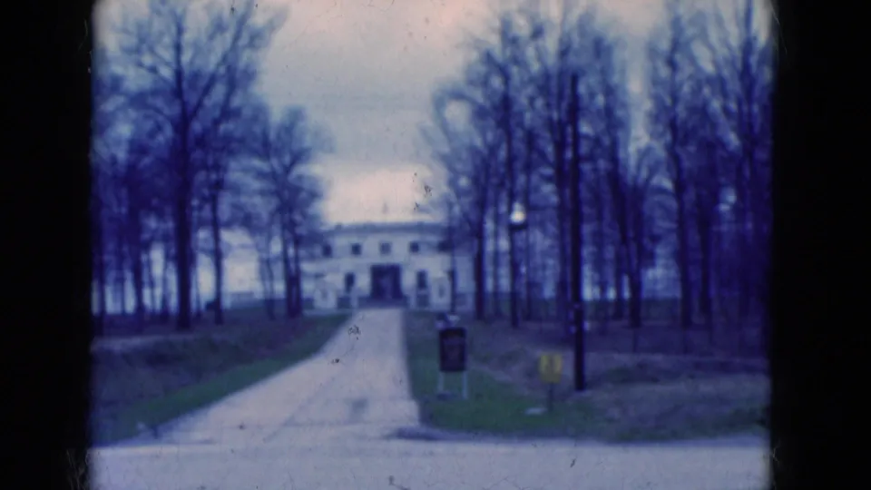1975 Florida 8mm frame of a white building approached by a driveway, with leafless trees, lawn, and signs near the foreground road.