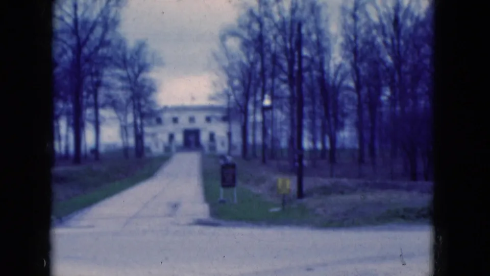 1975 Florida home-movie frame holding on a white building at the end of a drive, surrounded by bare trees and muted blue film color.