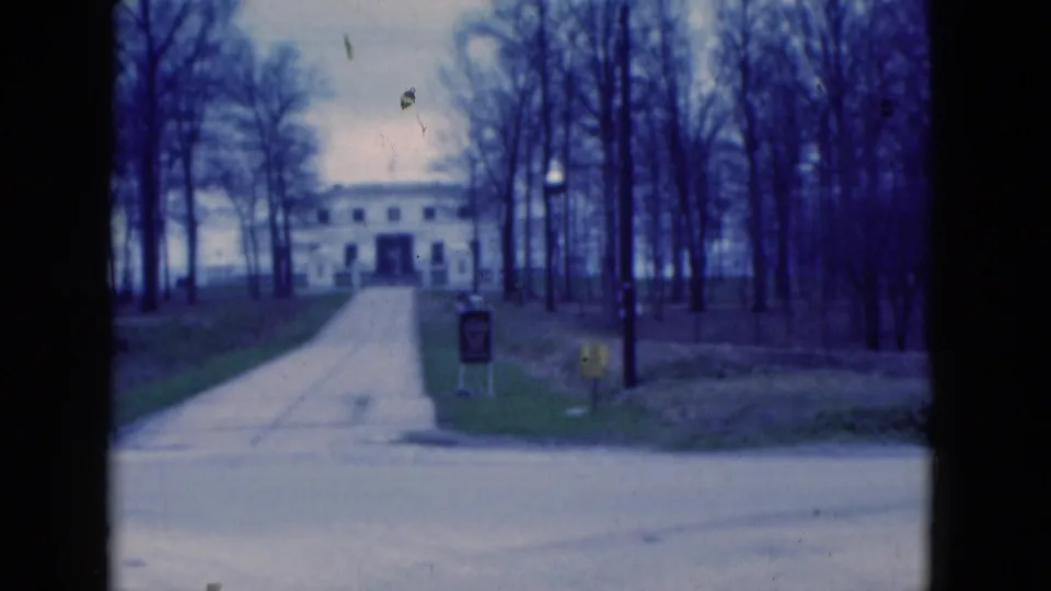 1975 Florida 8mm frame showing the same white building and driveway from a fixed position across the road, with signs and bare trees visible.