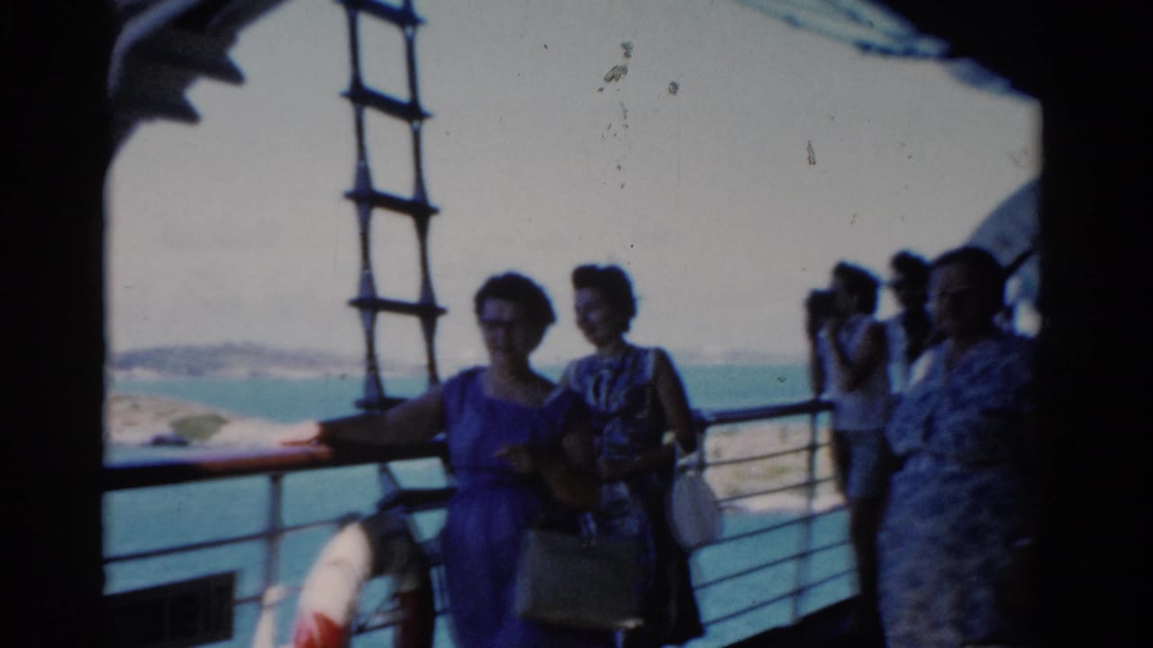 1975 Florida home-movie frame of women in summer dresses standing on a boat deck near a railing, with blue water and distant coast behind.