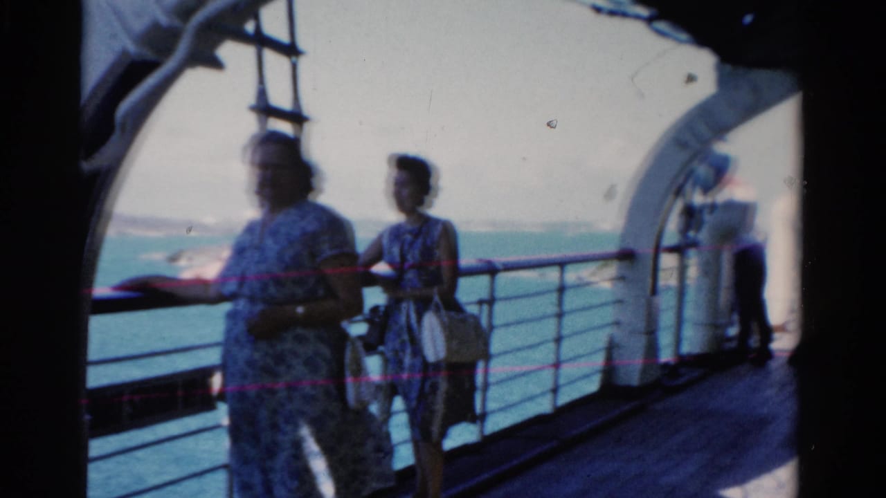 Women passengers stand along a white railing with water and shoreline behind them.
