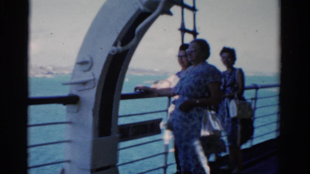 In 1975 Florida home-movie footage, three passengers stand on a ship deck near a white archway and look across the water.