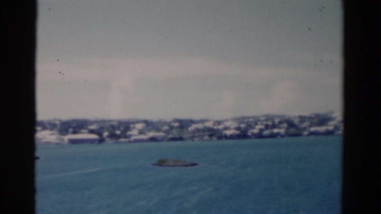 1975 Florida coastal view from the water, with pale shoreline buildings and a small rocky islet beneath a hazy sky.