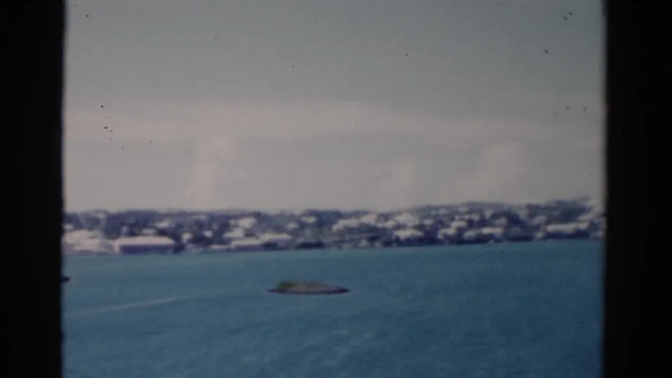 1975 Florida coastal view from the water, with pale shoreline buildings and a small rocky islet beneath a hazy sky.