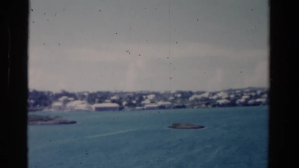 1975 Florida 8mm frame with blue-green water, low rocky islets, and a hazy shoreline with pale buildings in the distance.