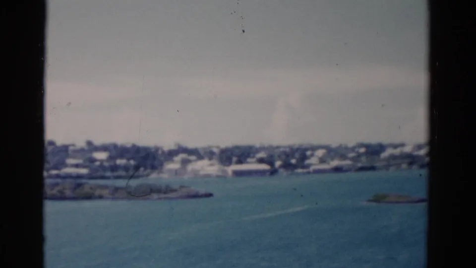 1975 Florida coastal view through vessel window bars, with calm blue water, hazy sky, and a distant low-rise shoreline.