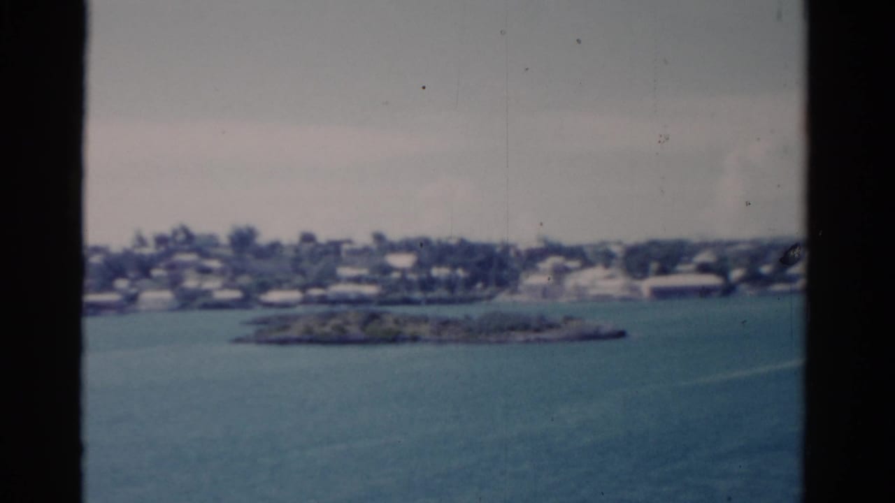 1975 Florida shoreline viewed from a vessel, with blue water, a small landform, low buildings, trees, and dark window bars.