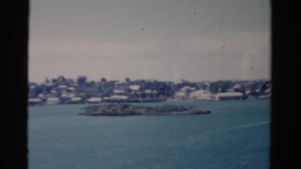 1975 Florida shoreline viewed from a vessel, with blue water, a small landform, low buildings, trees, and dark window bars.