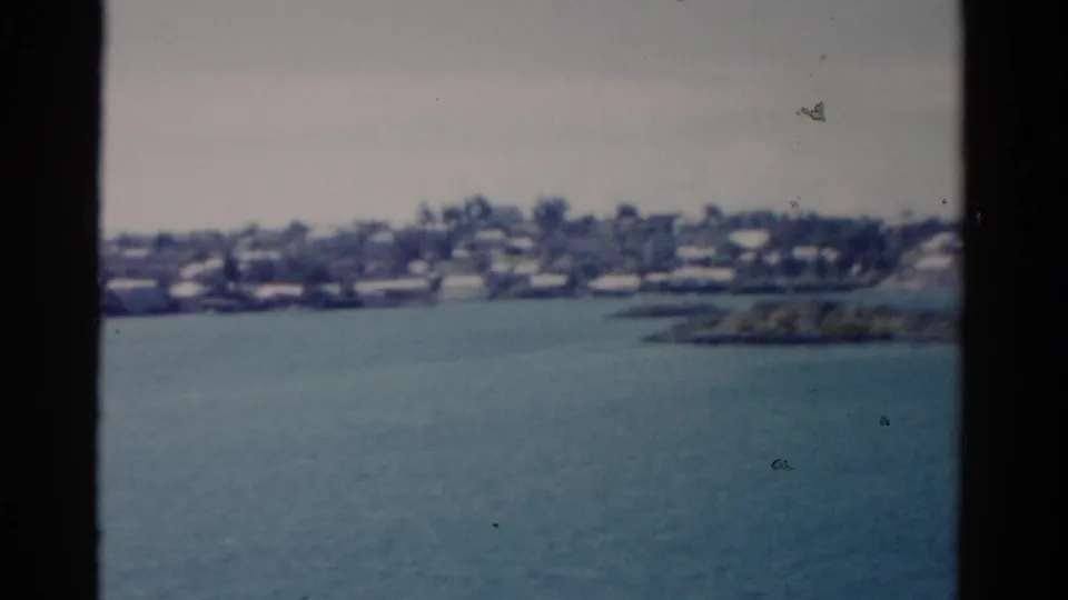 Soft 8mm view of a Florida waterfront in 1975, with blue water, trees, low buildings, and dark vertical window framing.