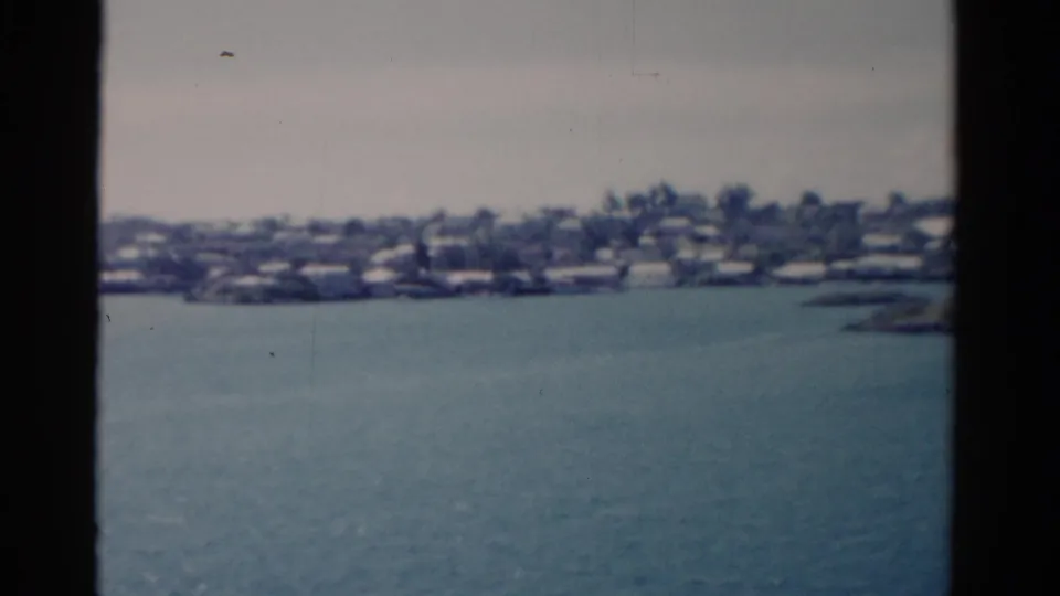 1975 Florida coastal water view with a distant tree-lined shoreline, low buildings, pale sky, and 8mm film grain.