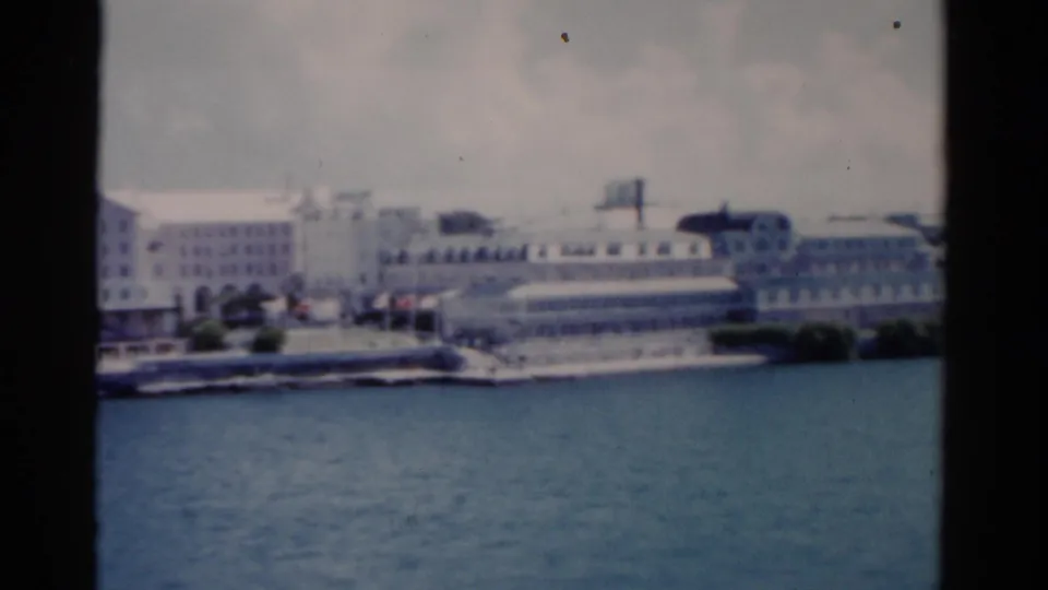 1975 Florida 8mm frame with waterfront buildings and a low shoreline complex seen from across blue water.