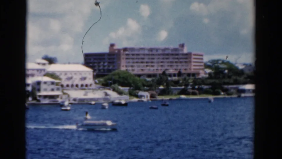1975 Florida waterfront view with a passing motorboat, anchored boats, white buildings, and a large hotel beyond the water.
