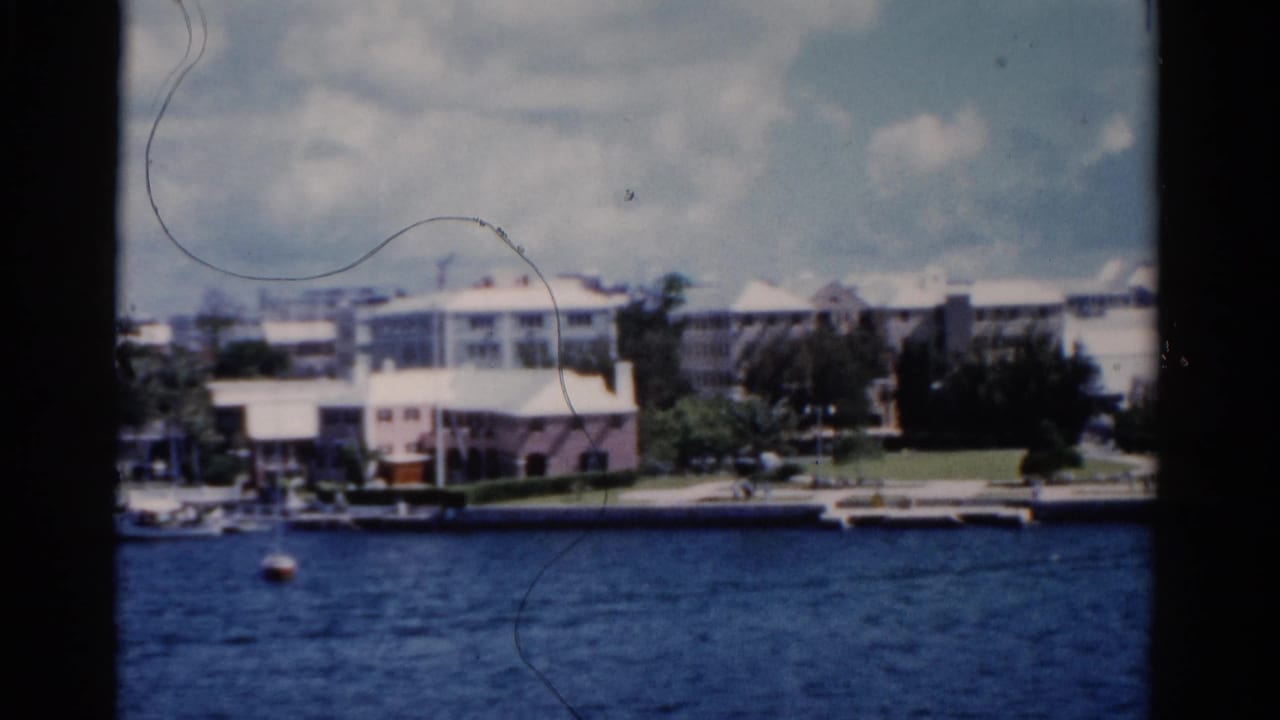 1975 Florida 8mm frame showing pastel waterfront buildings, trees, a seawall, blue water, and scattered clouds.