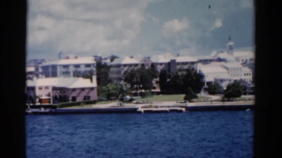 1975 Florida 8mm still of a sunny waterfront, with blue water, low-rise buildings, trees, lawns, and distant white roofs.