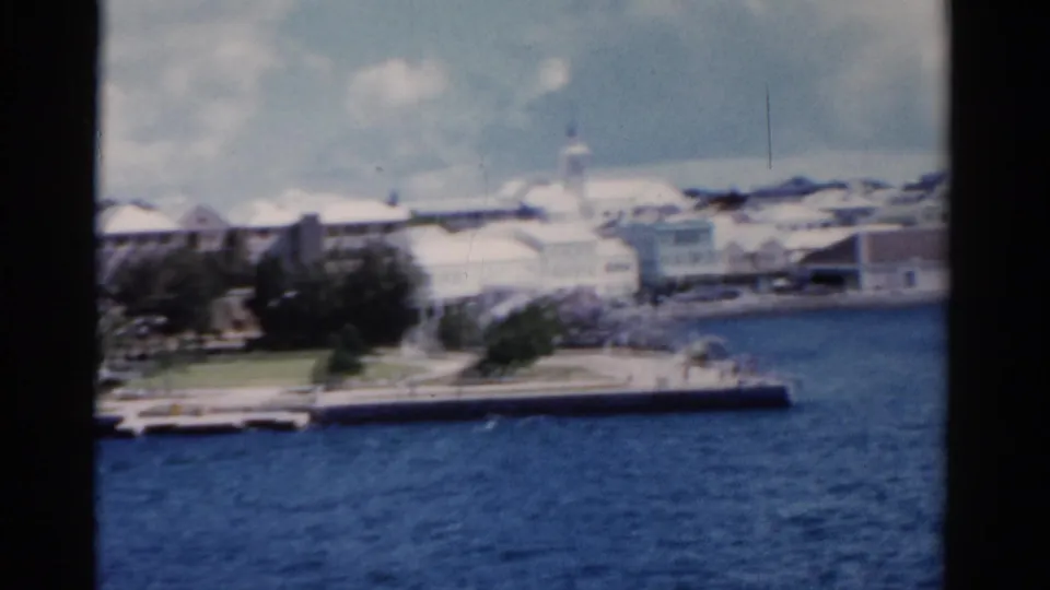 Soft 1975 Florida coastal view showing a park, promenade, pale buildings, water, and bright clouded sky.