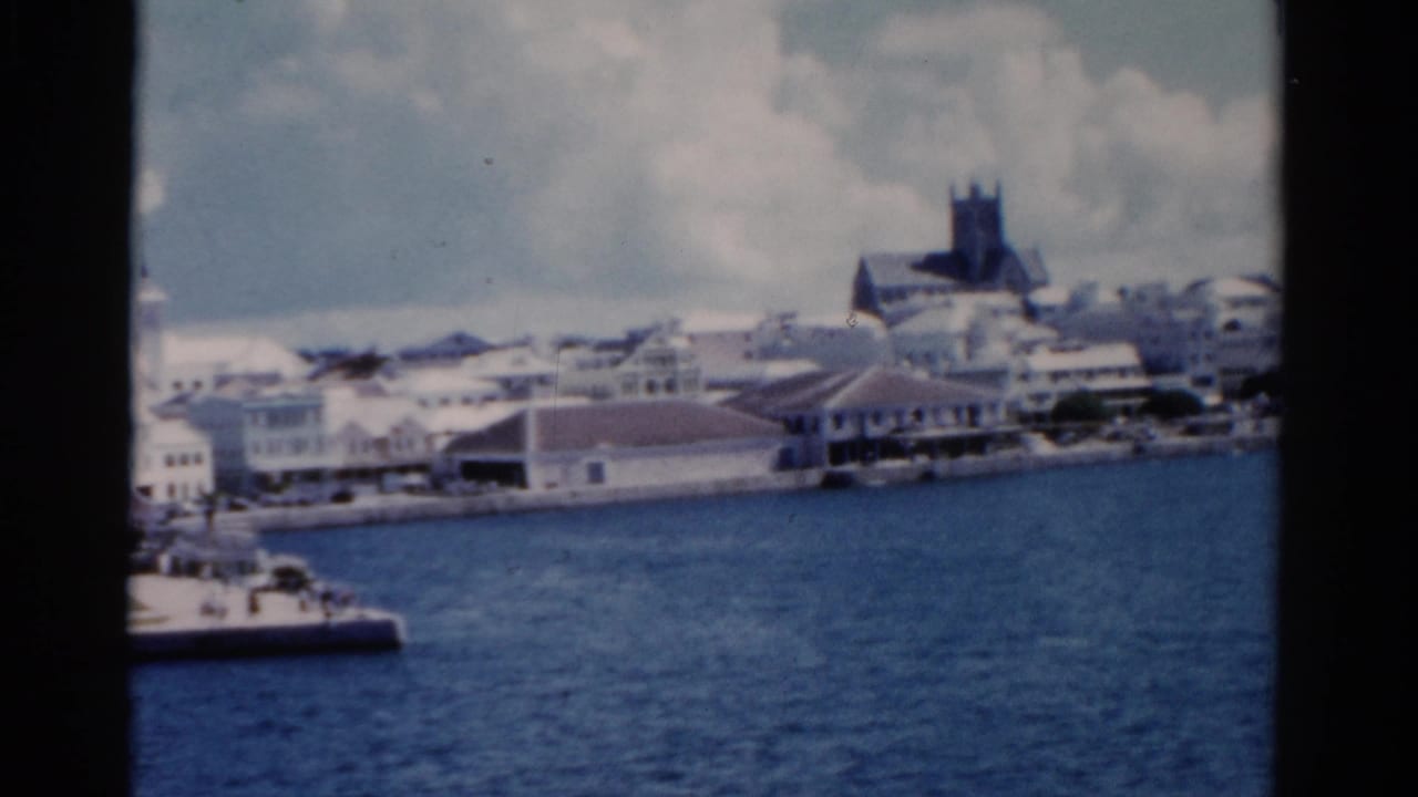 Harbor shoreline with white buildings, docks, and a church tower under a cloudy sky.