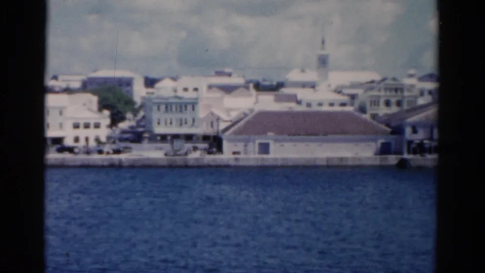 1975 Florida waterfront scene on 8mm with rippled water, pale buildings, and a distant spire under clouds.