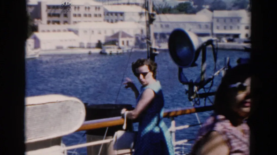 On a 1975 Florida boat deck, a woman in a blue dress looks toward the camera as another passenger passes in the foreground.