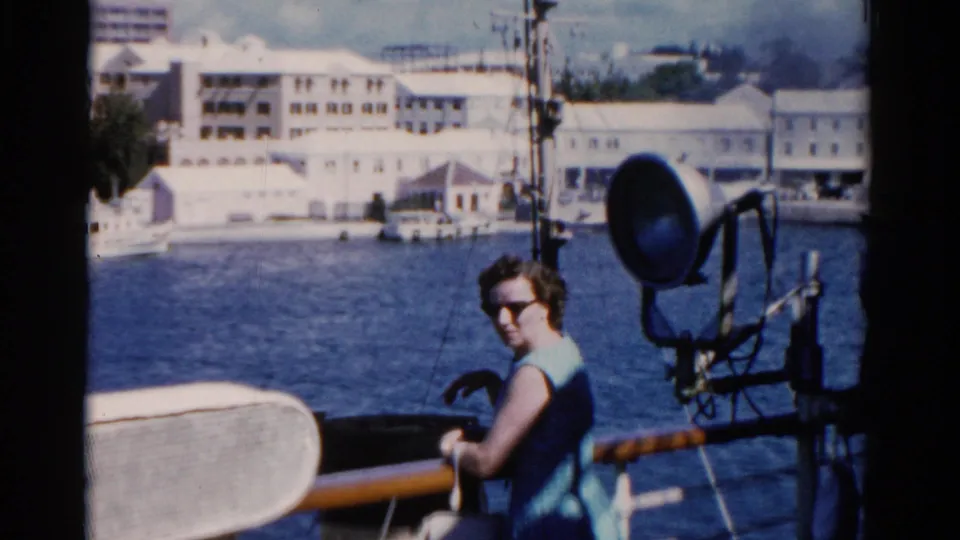 1975 Florida boat-deck view with a woman in a blue dress beside railings, deck equipment, blue water, and shoreline buildings.