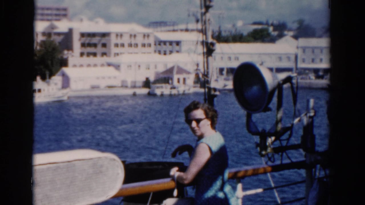 Woman in sunglasses stands by a ship railing with waterfront buildings across the water.