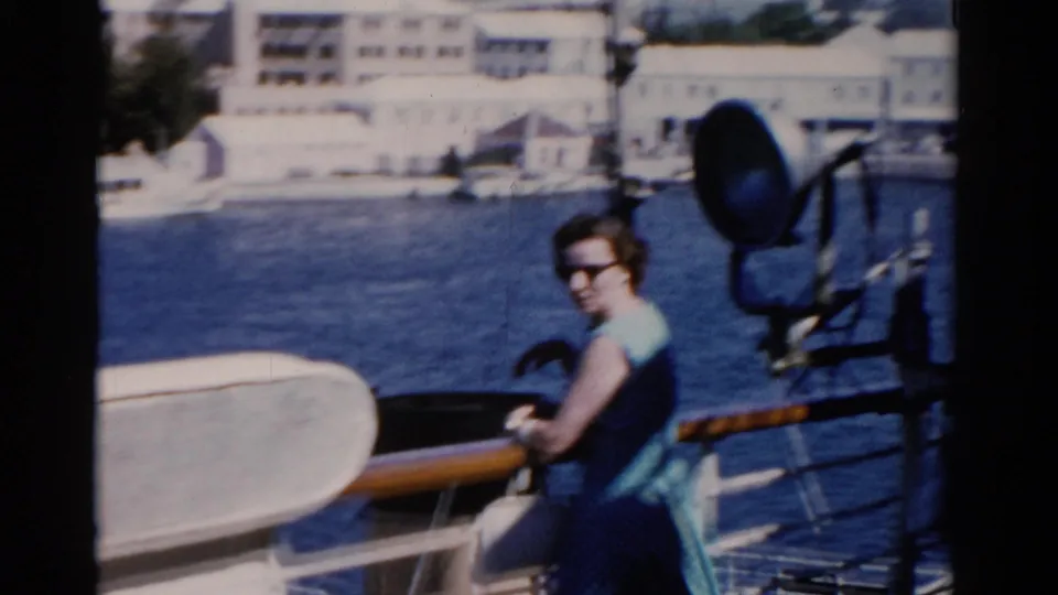 Woman in sunglasses on a Florida ship deck, with a large spotlight, wooden rail, water, and pale waterfront buildings behind her.