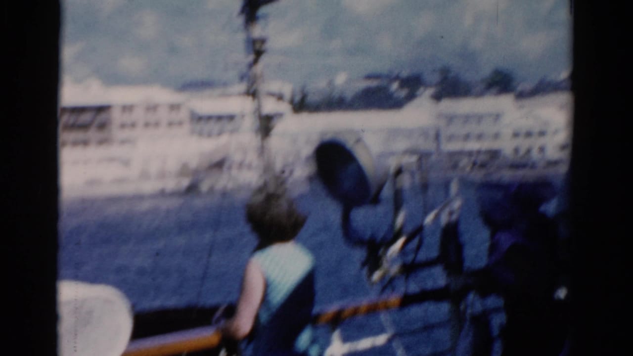1975 Florida home-movie frame showing a woman at a ship rail with water, white waterfront buildings, and a deck spotlight nearby.