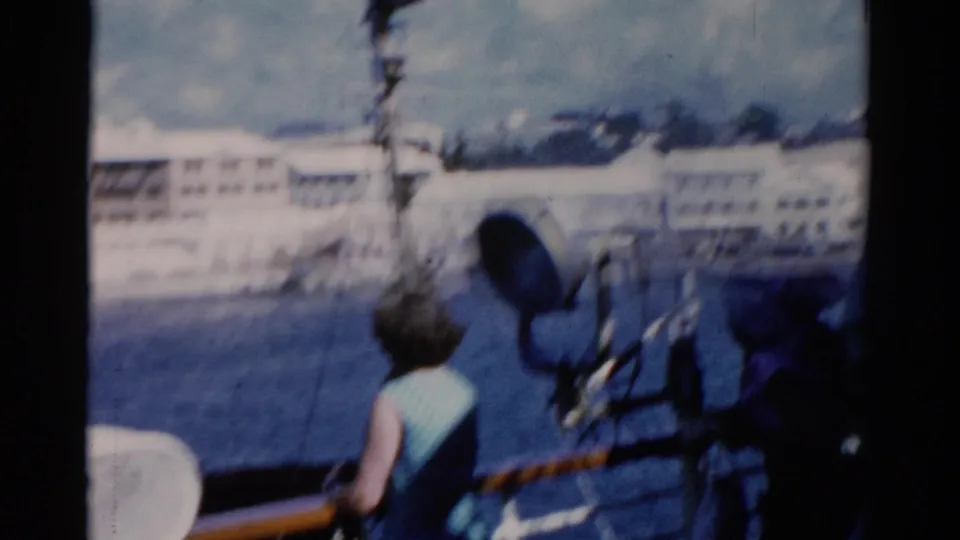 1975 Florida home-movie frame showing a woman at a ship rail with water, white waterfront buildings, and a deck spotlight nearby.