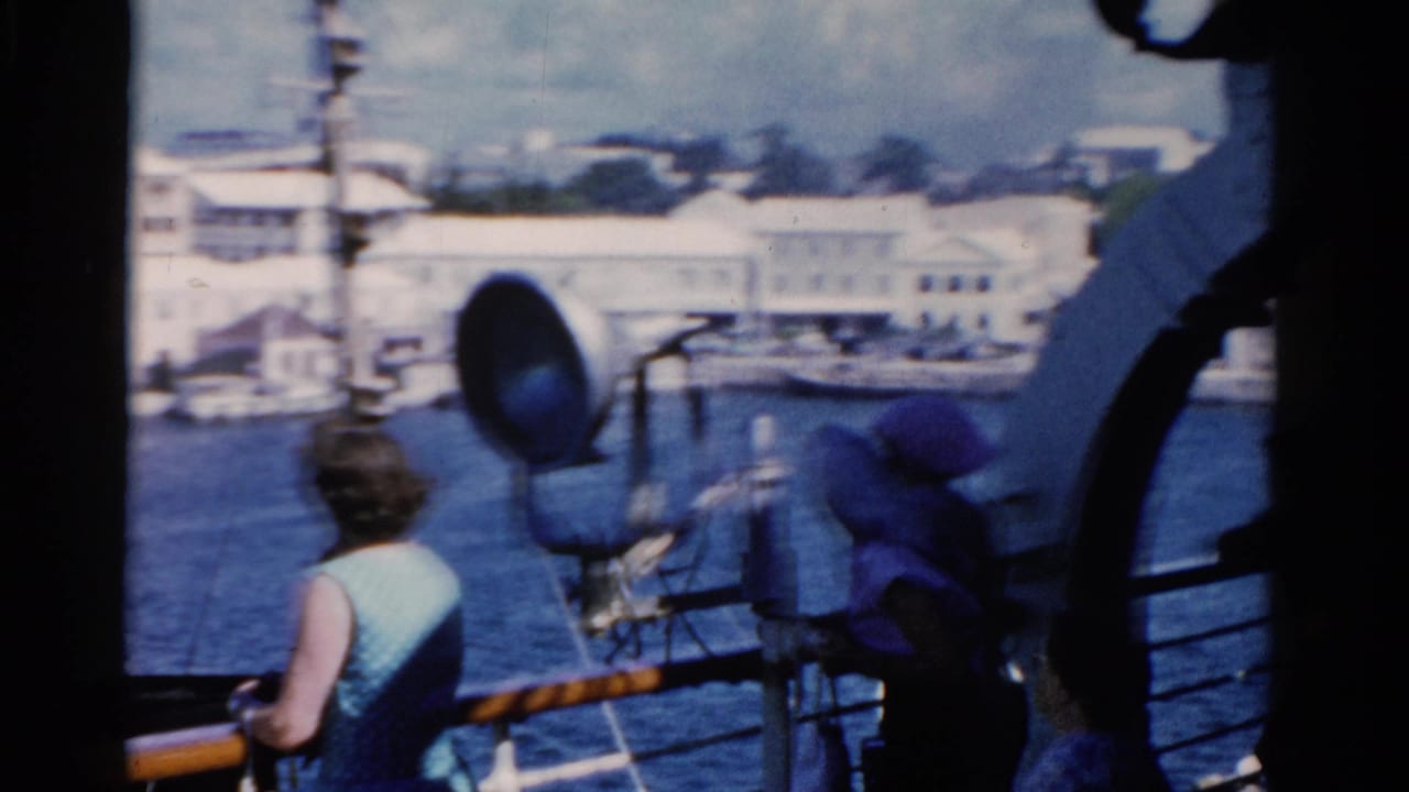 Ship deck view with woman at the rail, spotlight fixture, water, and waterfront buildings.
