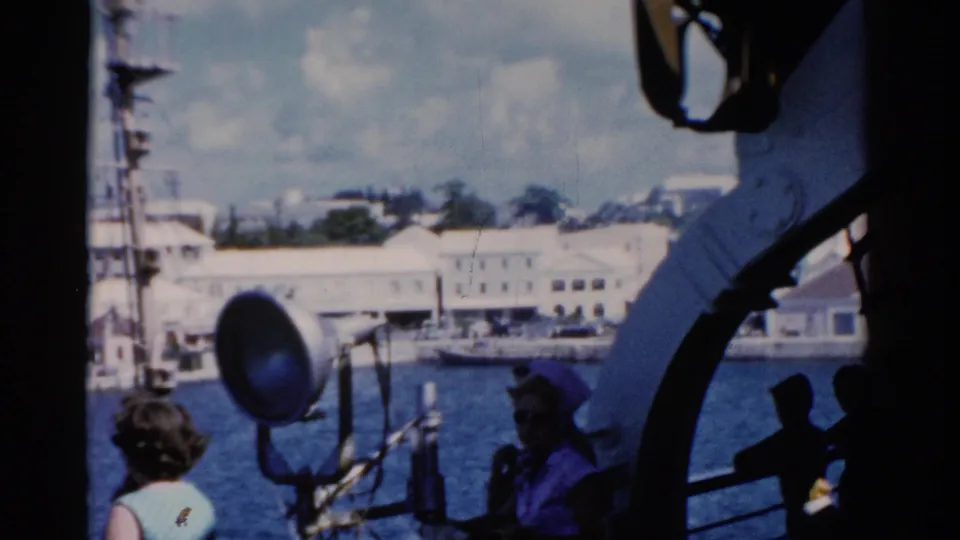 1975 Florida ship-deck frame showing the harbor through deck equipment, with the blue-dressed woman and another passenger visible.