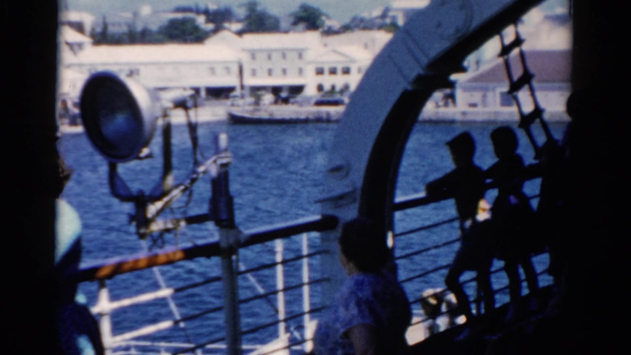 Shipboard view in 1975 Florida with passengers near the rail, a large spotlight, blue water, and waterfront buildings beyond.