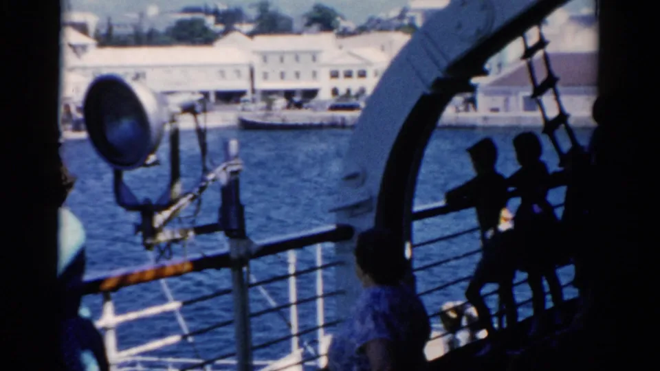 Shipboard view in 1975 Florida with passengers near the rail, a large spotlight, blue water, and waterfront buildings beyond.