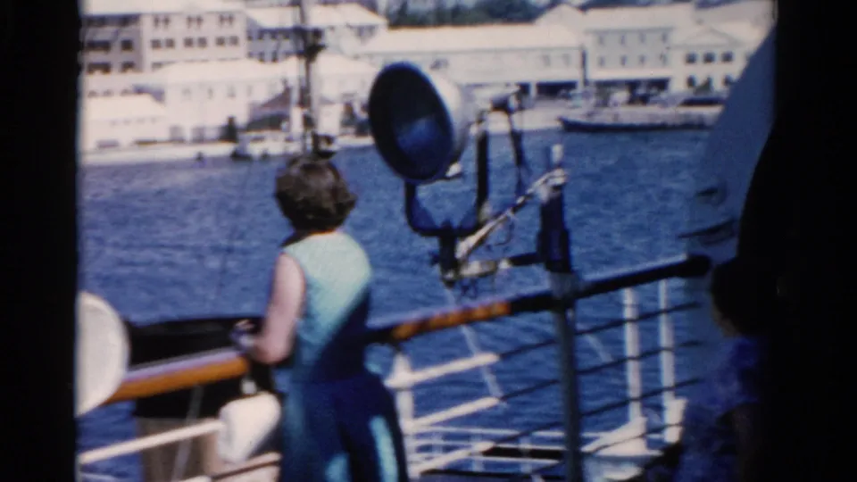 1975 Florida deck view with a woman in a light blue dress at the railing, harbor water, and white buildings across the shore.