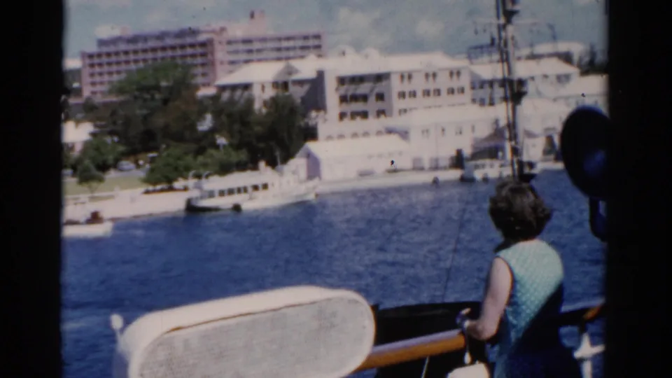 1975 Florida home-movie frame with ship deck equipment in front and a woman watching a harbor shoreline with boats and buildings.