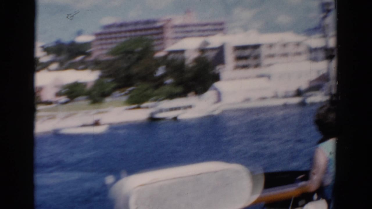1975 Florida 8mm frame looking from a boat toward white resort buildings across blue water, with a passenger partly visible at right.