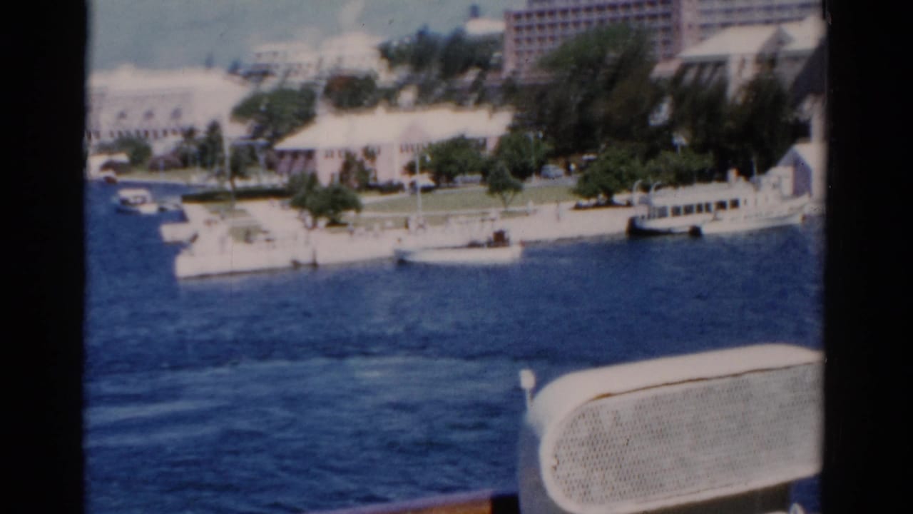 1975 Florida 8mm view across blue water toward white resort buildings, trees, docks, and a foreground boat seat or rail.