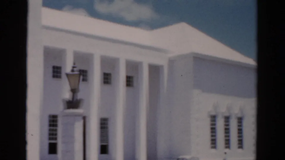1975 Florida 8mm frame of a white institutional-looking building facade, with columns, windows, lamppost, and roofline under blue sky.
