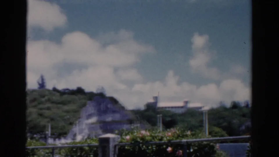 1975 Florida 8mm closing frame of a green hillside, shrubs, railing, and distant white structure through a window opening.