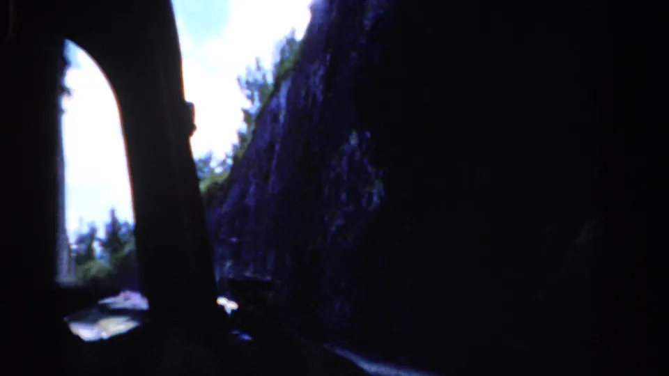 A dark in-car view frames a bright gap between rock faces, with trees and road surface visible beyond in 1970s 8mm footage.