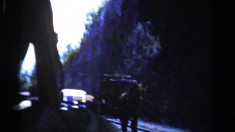 The moving viewpoint looks down a narrow road bordered by rock walls, with distant vehicles and a carriage-like silhouette in shadow.