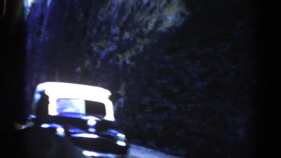 A bright-roofed vehicle passes near a dark rock wall and trees, rendered with strong blue shadows in the 8mm scan.