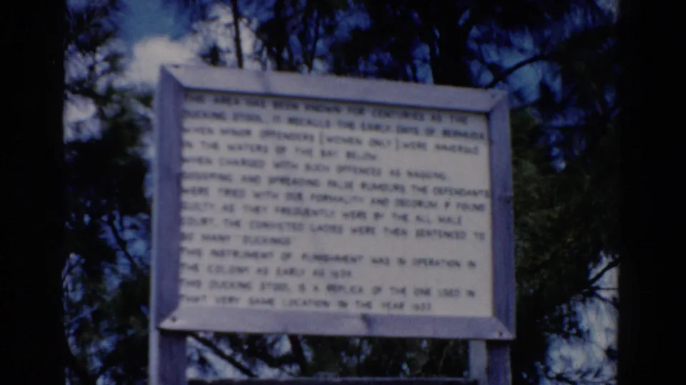 1975 8mm view of the historical marker, with its text more prominent but still soft, set against pine trees.