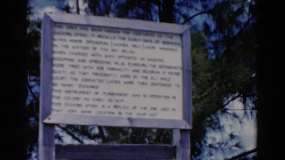 1975 Florida 8mm frame showing the same historical sign, softly focused with saturated blue sky and green pine foliage behind it.