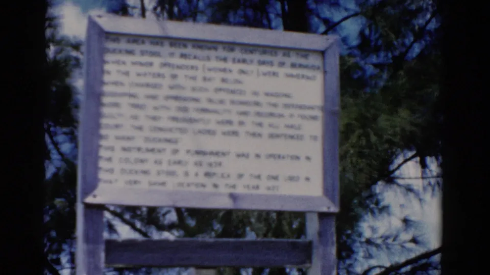 1975 8mm Florida frame of a wooden historical sign, partly soft in focus, with dark tree trunks and green foliage behind it.