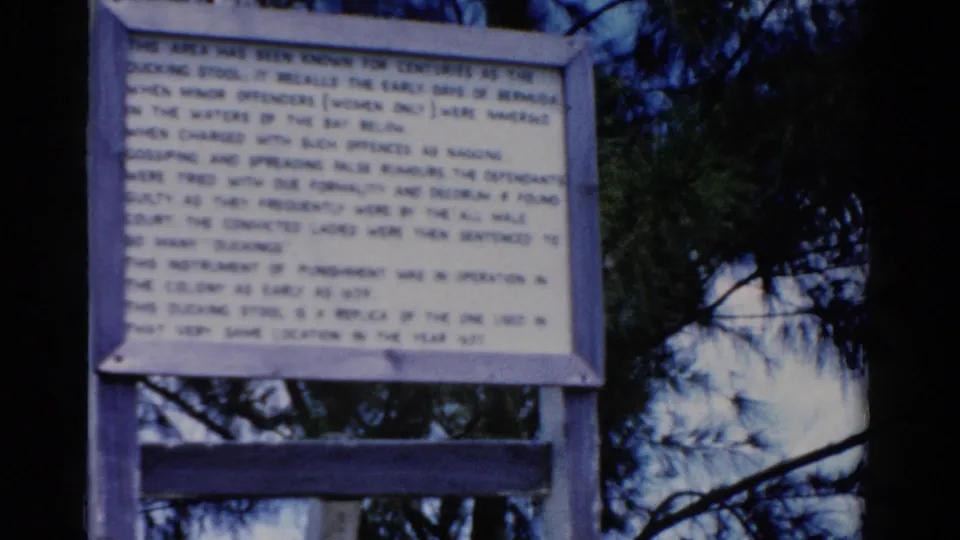 The camera holds on a text-heavy outdoor sign with trees around it, maintaining the home-movie travel perspective.