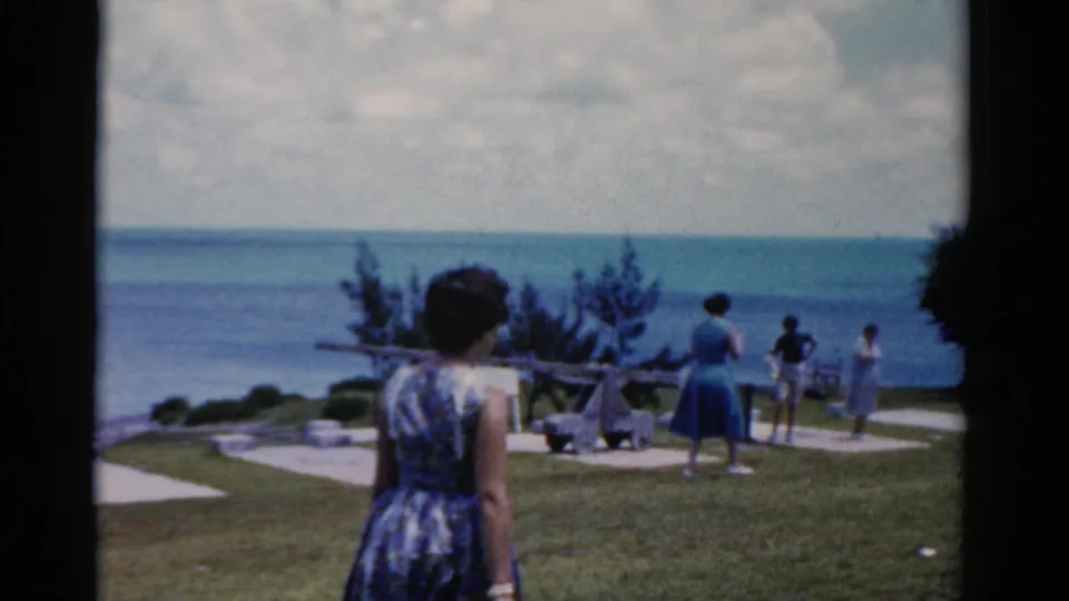 1975 Florida home-movie frame showing women in dresses on a grassy seaside overlook with the ocean beyond.