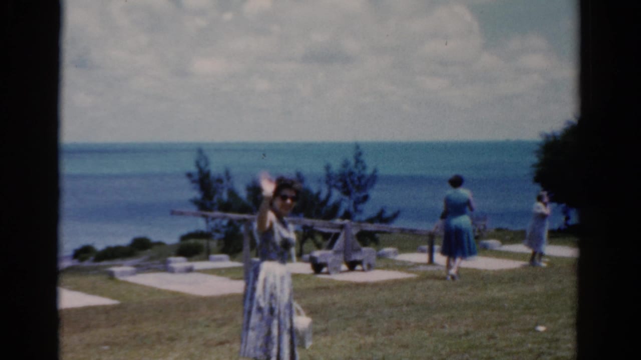 Woman in sunglasses waves on a grassy seaside overlook with other women near the water.